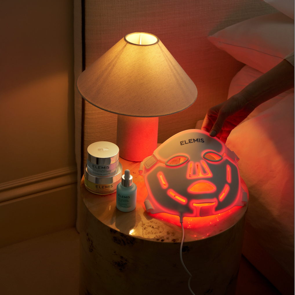 A person's hand is adjusting an ELEMIS LED light therapy face mask on a bedside table next to a lit lamp and skincare products.