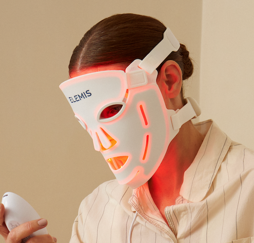 A person wearing a glowing white LED facial therapy mask and holding a matching handheld device, seated indoors against a plain beige wall.