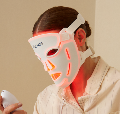 A person wearing a glowing white LED facial therapy mask and holding a matching handheld device, seated indoors against a plain beige wall.