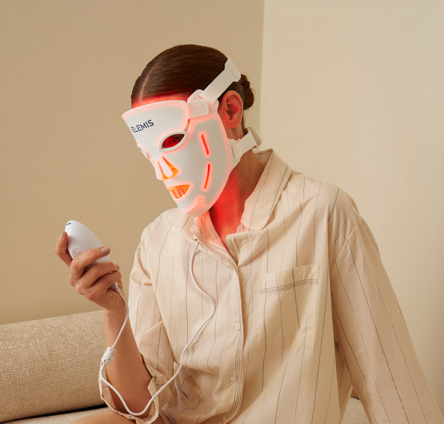 A person wearing a glowing white LED facial therapy mask and holding a matching handheld device, seated indoors against a plain beige wall.