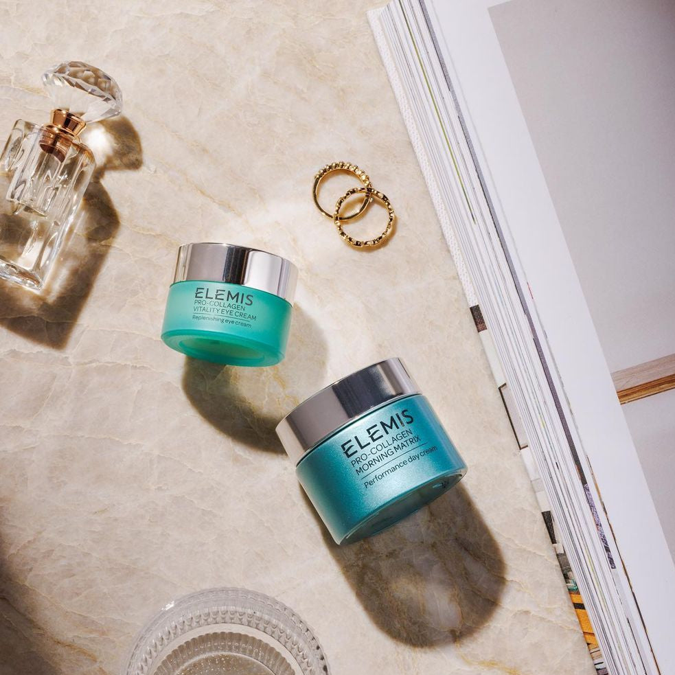 Two Elemis skincare jars on a marble surface with a magazine and perfume bottle in the background.
