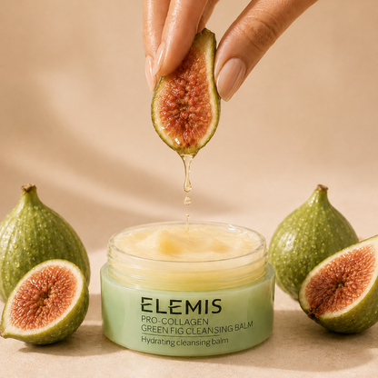 Elemis Green Fig Cleansing Balm in a light green jar, with a hand holding a sliced fig above it as honey drips into the balm. More whole and sliced figs are arranged around the jar on a warm beige background.