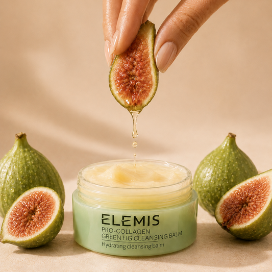 Elemis Green Fig Cleansing Balm in a light green jar, with a hand holding a sliced fig above it as honey drips into the balm. More whole and sliced figs are arranged around the jar on a warm beige background.